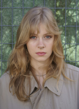 Eine junge Frau mit langen blonden Haaren und einem Nasenring steht vor einem grünen Maschendrahtzaun, trägt einen beigen Mantel und blickt mit neutralem Gesichtsausdruck direkt in die Kamera. 