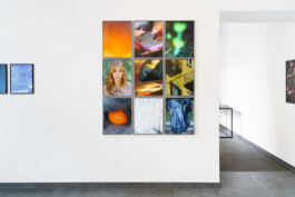 An einer weißen Galeriewand sind neun quadratische Fotografien zu sehen, die in einem Raster angeordnet sind. Sie zeigen abstrakte Motive, Porträts und urbane Szenen. Der Raum hat einen grauen Fliesenboden und eine Tür, die zu einem weiteren Raum führt. 