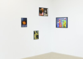 An einer weißen Eckwand hängen vier gerahmte Fotografien. Zu sehen sind bunte Reinigungsmittel, ein orangefarbener Luftballon, eine gelbe Treppe und eine Nahaufnahme von Händen neben einem Tisch. Der Raum hat einen hellen Boden. 