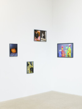 Vier gerahmte Fotografien hängen an einer weißen Galeriewand. Zu den Motiven gehören ein gelber Ballon, bunte Getränke, ein gelbes Gebäude mit Treppen und eine Nahaufnahme von Blumen mit Vögeln. Der Boden ist hell. 