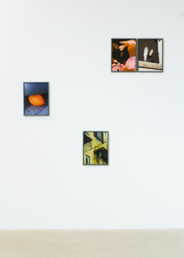 An einer weißen Wand hängen vier gerahmte Fotografien. Die Bilder zeigen einen orangefarbenen Luftballon, zwei Nahaufnahmen von Objekten (darunter eine Feder) und eine gelbe Gebäudefassade. Der Boden darunter ist schlicht und beige. 