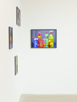 An einer weißen Galeriewand hängen vier gerahmte Kunstwerke. Links sind drei kleinere Bilder zu sehen, während das größte, das nebeneinander präsentierte bunte Reinigungsprodukte und Snacks zeigt und prominent rechts hängt. 